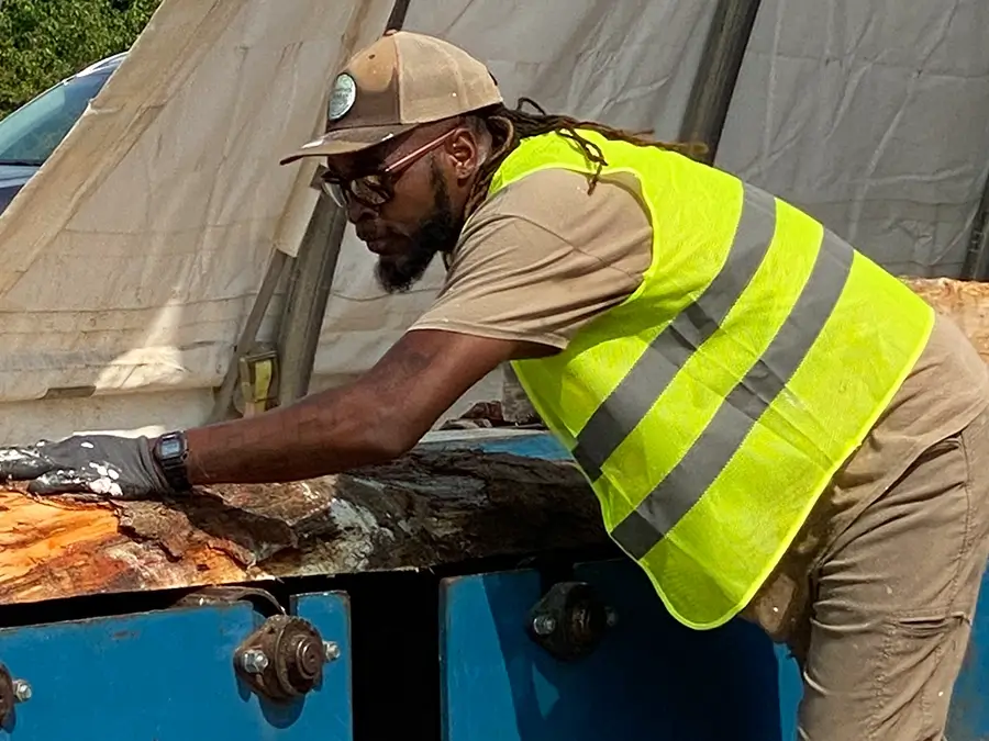 Man in an outdoor wood processing yard feeding a section of a log through a planing machine.