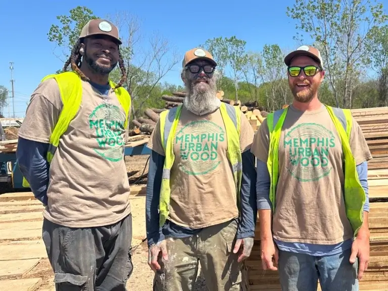 Three men with construction vests in a wood processing yard. They're smiling and wearing Memphis Urban Wood t-shirts.
