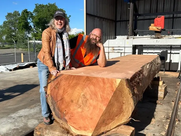 Caucasian woman and man in construction vest outdoors laughing and leaning on a very large tree log that's being processed into usable wood.