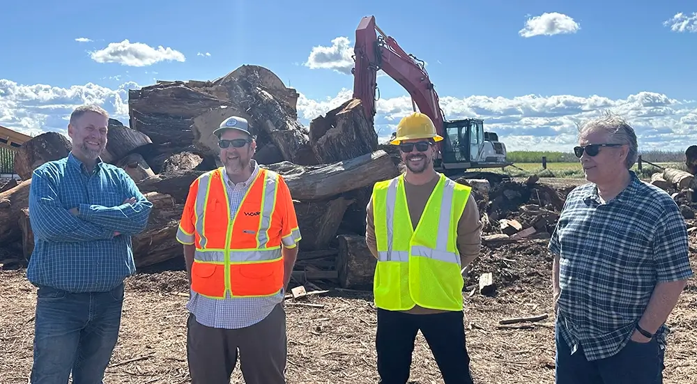 Four adult men, two with hard hats and yellow construction vests, stand outdoors smiling in front of a pile of large tree logs and a backhoe.